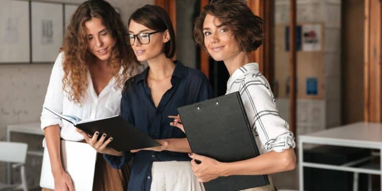 Três jovens mulheres profissionais em um escritório moderno; uma à direita segura uma prancheta e olha para a câmera sorrindo, enquanto as outras duas ao centro e à esquerda consultam juntas um notebook e uma pasta.