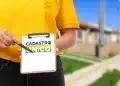 Close de uma pessoa vestindo camiseta amarela e calça jeans, segurando uma prancheta de madeira com uma folha branca. Na folha, destaca-se o logotipo do Cadastro Único em azul, amarelo e verde. A pessoa aponta para o logotipo com uma caneta preta. Ao fundo, uma área residencial com casas populares desfocadas.