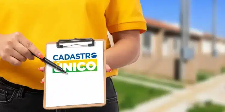 Close de uma pessoa vestindo camiseta amarela e calça jeans, segurando uma prancheta de madeira com uma folha branca. Na folha, destaca-se o logotipo do Cadastro Único em azul, amarelo e verde. A pessoa aponta para o logotipo com uma caneta preta. Ao fundo, uma área residencial com casas populares desfocadas.