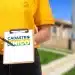 Close de uma pessoa vestindo camiseta amarela e calça jeans, segurando uma prancheta de madeira com uma folha branca. Na folha, destaca-se o logotipo do Cadastro Único em azul, amarelo e verde. A pessoa aponta para o logotipo com uma caneta preta. Ao fundo, uma área residencial com casas populares desfocadas.