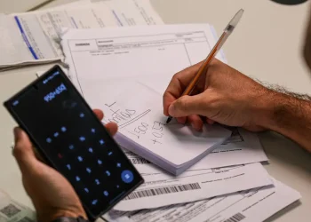 Mutirão de dívidas bancárias 2026 — homem segura celular com calculadora aberta enquanto anota valores em bloco de notas sobre uma mesa com boletos e contas a pagar, representando o planejamento financeiro para renegociar débitos durante o mutirão da Febraban