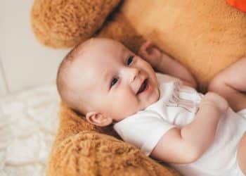 Bebê sorridente deitado de costas em um urso de pelúcia gigante, representando a doçura dos nomes clássicos que são tendência em 2026.