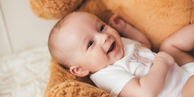 Bebê sorridente deitado de costas em um urso de pelúcia gigante, representando a doçura dos nomes clássicos que são tendência em 2026.