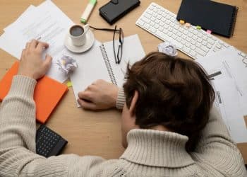 Pessoa com a cabeça apoiada na mesa, cercada por papéis e xícaras, simbolizando a procrastinação e o acúmulo de tarefas.