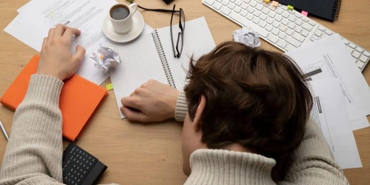 Pessoa com a cabeça apoiada na mesa, cercada por papéis e xícaras, simbolizando a procrastinação e o acúmulo de tarefas.