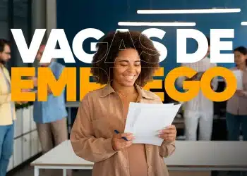 Uma mulher negra de cabelo afro, vestindo uma camisa bege, sorrindo e segurando e lendo um papel e uma caneta azul na mão, em um ambiente de escritório com outras pessoas desfocadas ao fundo. O texto "VAGAS DE EMPREGO" está sobreposto em branco e laranja na parte superior da imagem.