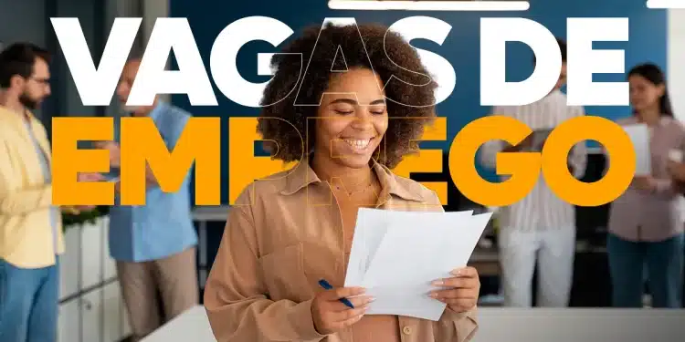 Uma mulher negra de cabelo afro, vestindo uma camisa bege, sorrindo e segurando e lendo um papel e uma caneta azul na mão, em um ambiente de escritório com outras pessoas desfocadas ao fundo. O texto "VAGAS DE EMPREGO" está sobreposto em branco e laranja na parte superior da imagem.
