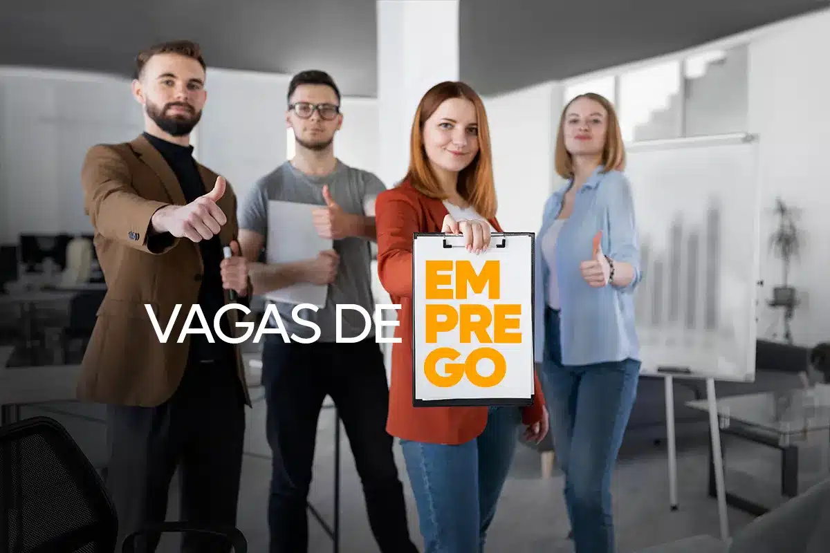 Grupo de quatro profissionais jovens em um escritório moderno fazendo sinal de positivo; a mulher ao centro segura uma prancheta com o texto "Vagas de Emprego" em laranja e branco.