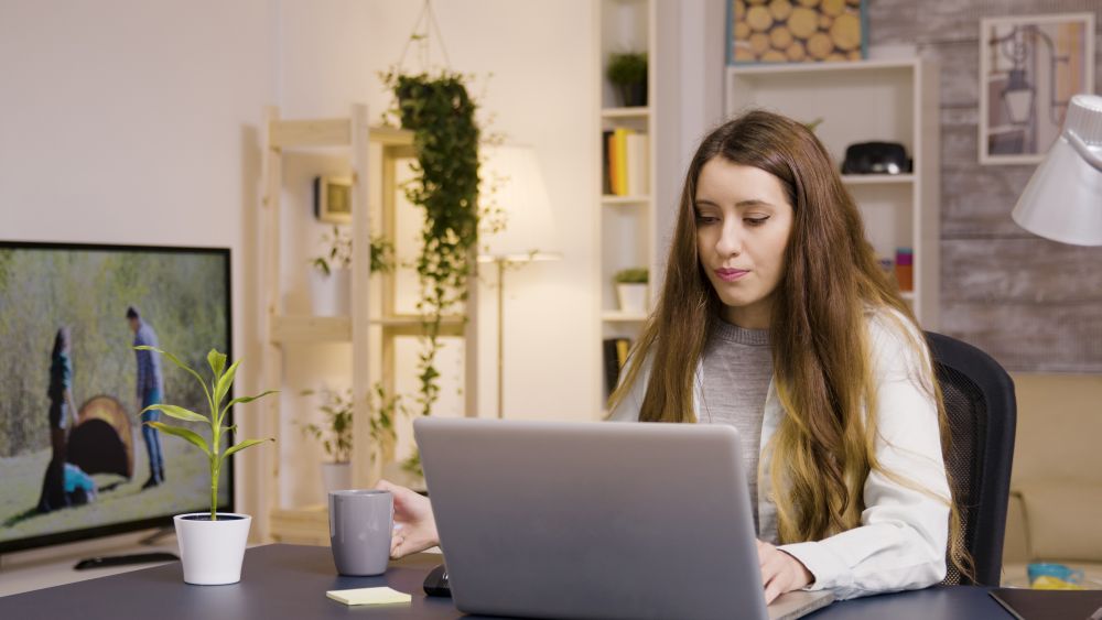 Mulher jovem de cabelos longos e castanhos trabalhando concentrada em um notebook prata em sua sala de estar decorada com plantas.