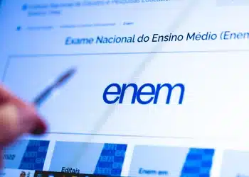 Tela do site do Enem exibida em computador com pessoa segurando caneta, representando orientações sobre a redação do exame.