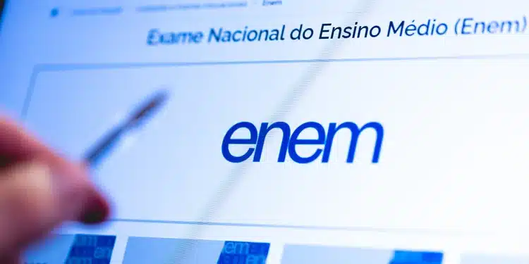Tela do site do Enem exibida em computador com pessoa segurando caneta, representando orientações sobre a redação do exame.