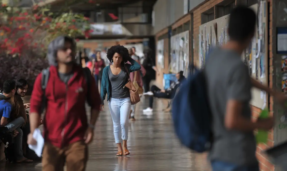 Estudante jovem negra com cabelo crespo caminhando pelo corredor externo de uma universidade federal carregando bolsa, com outros estudantes ao fundo em ambiente universitário arborizado, representando os beneficiários do programa Pé-de-Meia Licenciaturas 2026