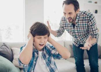 Pai gritando com o filho enquanto a criança tapa os ouvidos demonstrando sofrimento emocional dentro de casa.