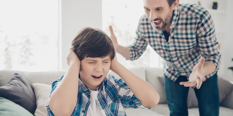 Pai gritando com o filho enquanto a criança tapa os ouvidos demonstrando sofrimento emocional dentro de casa.