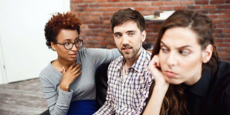 Três colegas de trabalho em um escritório durante uma pausa, com uma pessoa demonstrando desconforto enquanto as outras conversam.