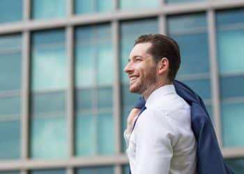 Homem jovem de perfil, sorrindo, veste camisa social branca e carrega um paletó azul sobre o ombro, com um edifício comercial moderno e espelhado ao fundo.