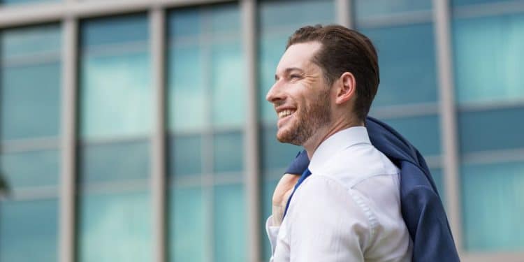 Homem jovem de perfil, sorrindo, veste camisa social branca e carrega um paletó azul sobre o ombro, com um edifício comercial moderno e espelhado ao fundo.