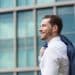 Homem jovem de perfil, sorrindo, veste camisa social branca e carrega um paletó azul sobre o ombro, com um edifício comercial moderno e espelhado ao fundo.