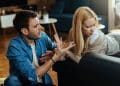 Homem em postura agressiva gesticulando com as mãos enquanto fala com uma mulher que desvia o rosto com expressão de desconforto e dúvida em uma sala de estar.