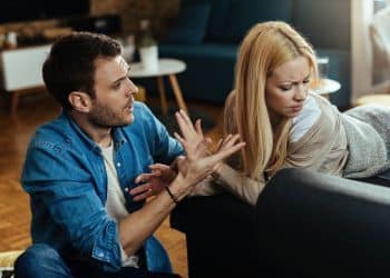 Homem em postura agressiva gesticulando com as mãos enquanto fala com uma mulher que desvia o rosto com expressão de desconforto e dúvida em uma sala de estar.