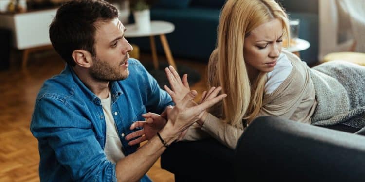 Homem em postura agressiva gesticulando com as mãos enquanto fala com uma mulher que desvia o rosto com expressão de desconforto e dúvida em uma sala de estar.