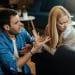 Homem em postura agressiva gesticulando com as mãos enquanto fala com uma mulher que desvia o rosto com expressão de desconforto e dúvida em uma sala de estar.