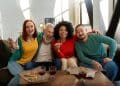 Um grupo diversificado de quatro amigos, de diferentes gerações e origens, sorrindo e se abraçando em um sofá. Uma mulher de cabelos ruivos faz um sinal de "V" com os dedos, um homem idoso de barba branca ri, uma mulher com cabelos cacheados segura uma fatia de pizza e um homem asiático sorri. À frente deles, uma mesa com pizza e copos com bebida. A imagem ilustra a conexão e a diversão em grupo.