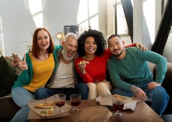 Um grupo diversificado de quatro amigos, de diferentes gerações e origens, sorrindo e se abraçando em um sofá. Uma mulher de cabelos ruivos faz um sinal de "V" com os dedos, um homem idoso de barba branca ri, uma mulher com cabelos cacheados segura uma fatia de pizza e um homem asiático sorri. À frente deles, uma mesa com pizza e copos com bebida. A imagem ilustra a conexão e a diversão em grupo.