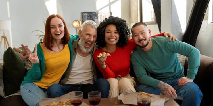 Um grupo diversificado de quatro amigos, de diferentes gerações e origens, sorrindo e se abraçando em um sofá. Uma mulher de cabelos ruivos faz um sinal de "V" com os dedos, um homem idoso de barba branca ri, uma mulher com cabelos cacheados segura uma fatia de pizza e um homem asiático sorri. À frente deles, uma mesa com pizza e copos com bebida. A imagem ilustra a conexão e a diversão em grupo.
