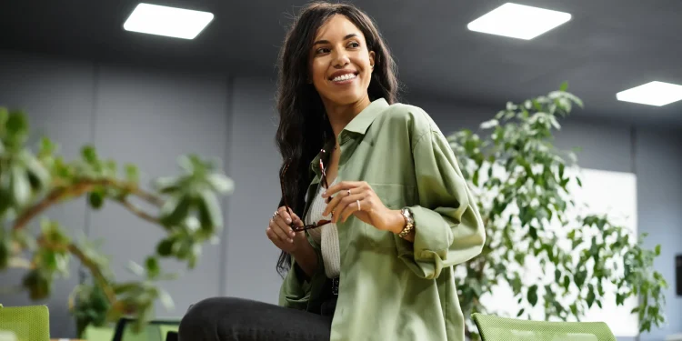 Mulher sorrindo com uma expressão confiante em ambiente de escritório, segurando óculos.