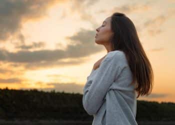 Mulher de perfil, com olhos fechados, refletindo ao ar livre, com o pôr do sol ao fundo, simbolizando força mental.