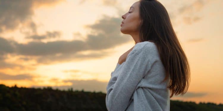 Mulher de perfil, com olhos fechados, refletindo ao ar livre, com o pôr do sol ao fundo, simbolizando força mental.