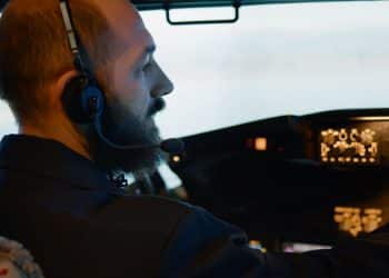 Piloto de aviação de perfil, com barba, usando fones de ouvido e microfone dentro de uma cabine de comando iluminada, operando o painel de controle de uma aeronave.