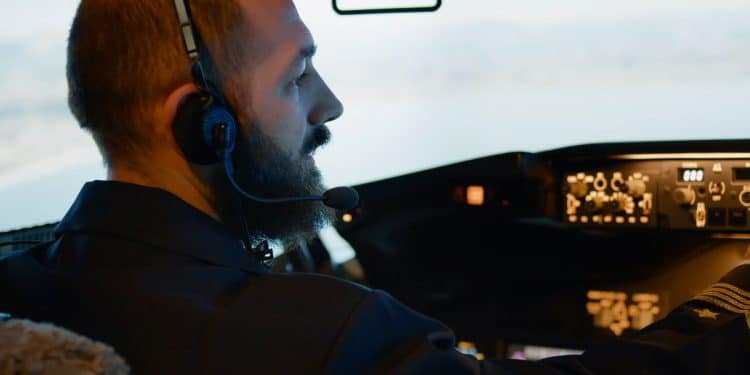 Piloto de aviação de perfil, com barba, usando fones de ouvido e microfone dentro de uma cabine de comando iluminada, operando o painel de controle de uma aeronave.