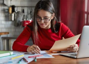 Jovem estudante com óculos revisando anotações e materiais sobre a mesa em preparação para o vestibular 2026.