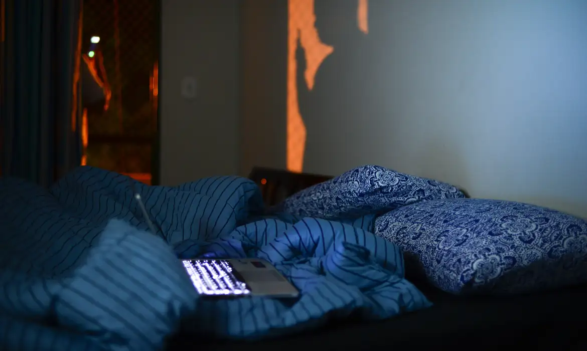 Cama desfeita à noite com smartphone iluminado entre os travesseiros e luz laranja entrando pela janela, representando o hábito de ficar acordado até tarde