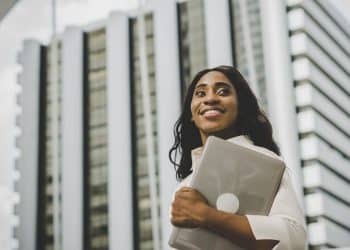 Mulher negra profissional, sorrindo e olhando para o alto, segurando livros de guias salariais. O livro à frente tem o texto: 'SALÁRIOS DE ATÉ R$ 50 MIL: VEJA AS PROFISSÕES QUE MAIS PAGAM EM CARGOS AVANÇADOS'. Ao fundo, edifícios de escritórios modernos. Foto de sucesso e carreira.