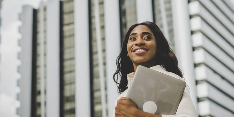 Mulher negra profissional, sorrindo e olhando para o alto, segurando livros de guias salariais. O livro à frente tem o texto: 'SALÁRIOS DE ATÉ R$ 50 MIL: VEJA AS PROFISSÕES QUE MAIS PAGAM EM CARGOS AVANÇADOS'. Ao fundo, edifícios de escritórios modernos. Foto de sucesso e carreira.