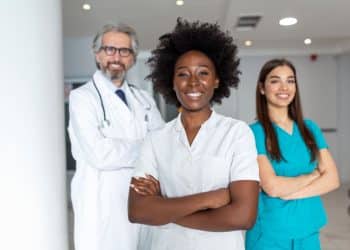 Fotografia de estúdio de uma equipe médica diversificada composta por três profissionais em pé, de frente para a câmera e sorrindo. No centro, uma jovem mulher negra com cabelos afro volumosos, vestindo um jaleco branco abotoado com os braços cruzados. À esquerda, ligeiramente recuado e desfocado, um homem grisalho de óculos e barba, vestindo jaleco branco com um estetoscópio no pescoço. À direita, desfocada, uma jovem mulher branca de cabelos longos castanhos, vestindo um esfoliante cirúrgico azul-turquesa com os braços cruzados. O fundo é um ambiente de clínica ou hospital com colunas brancas e luminárias de teto de LED retangulares sob um céu claro ao fundo.