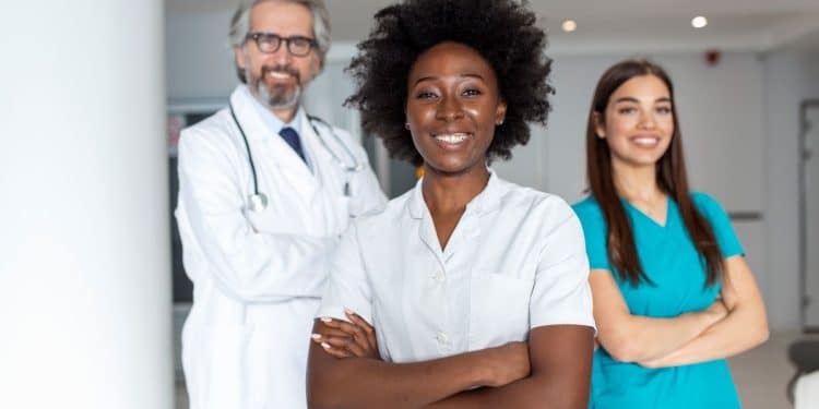 Fotografia de estúdio de uma equipe médica diversificada composta por três profissionais em pé, de frente para a câmera e sorrindo. No centro, uma jovem mulher negra com cabelos afro volumosos, vestindo um jaleco branco abotoado com os braços cruzados. À esquerda, ligeiramente recuado e desfocado, um homem grisalho de óculos e barba, vestindo jaleco branco com um estetoscópio no pescoço. À direita, desfocada, uma jovem mulher branca de cabelos longos castanhos, vestindo um esfoliante cirúrgico azul-turquesa com os braços cruzados. O fundo é um ambiente de clínica ou hospital com colunas brancas e luminárias de teto de LED retangulares sob um céu claro ao fundo.