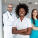 Fotografia de estúdio de uma equipe médica diversificada composta por três profissionais em pé, de frente para a câmera e sorrindo. No centro, uma jovem mulher negra com cabelos afro volumosos, vestindo um jaleco branco abotoado com os braços cruzados. À esquerda, ligeiramente recuado e desfocado, um homem grisalho de óculos e barba, vestindo jaleco branco com um estetoscópio no pescoço. À direita, desfocada, uma jovem mulher branca de cabelos longos castanhos, vestindo um esfoliante cirúrgico azul-turquesa com os braços cruzados. O fundo é um ambiente de clínica ou hospital com colunas brancas e luminárias de teto de LED retangulares sob um céu claro ao fundo.