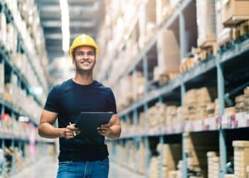 Profissional sorridente usando capacete de segurança, segurando uma prancheta em ambiente de armazém, representando carreiras de nível médio em alta.