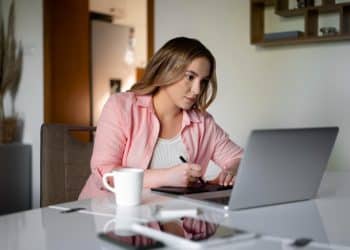 Mulher de camisa rosa concentrada usando mesa digitalizadora e notebook em ambiente doméstico com xícara de café à frente