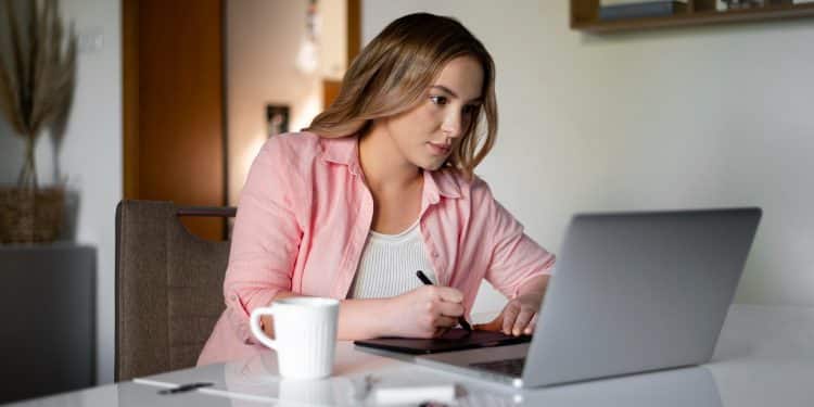 Mulher de camisa rosa concentrada usando mesa digitalizadora e notebook em ambiente doméstico com xícara de café à frente