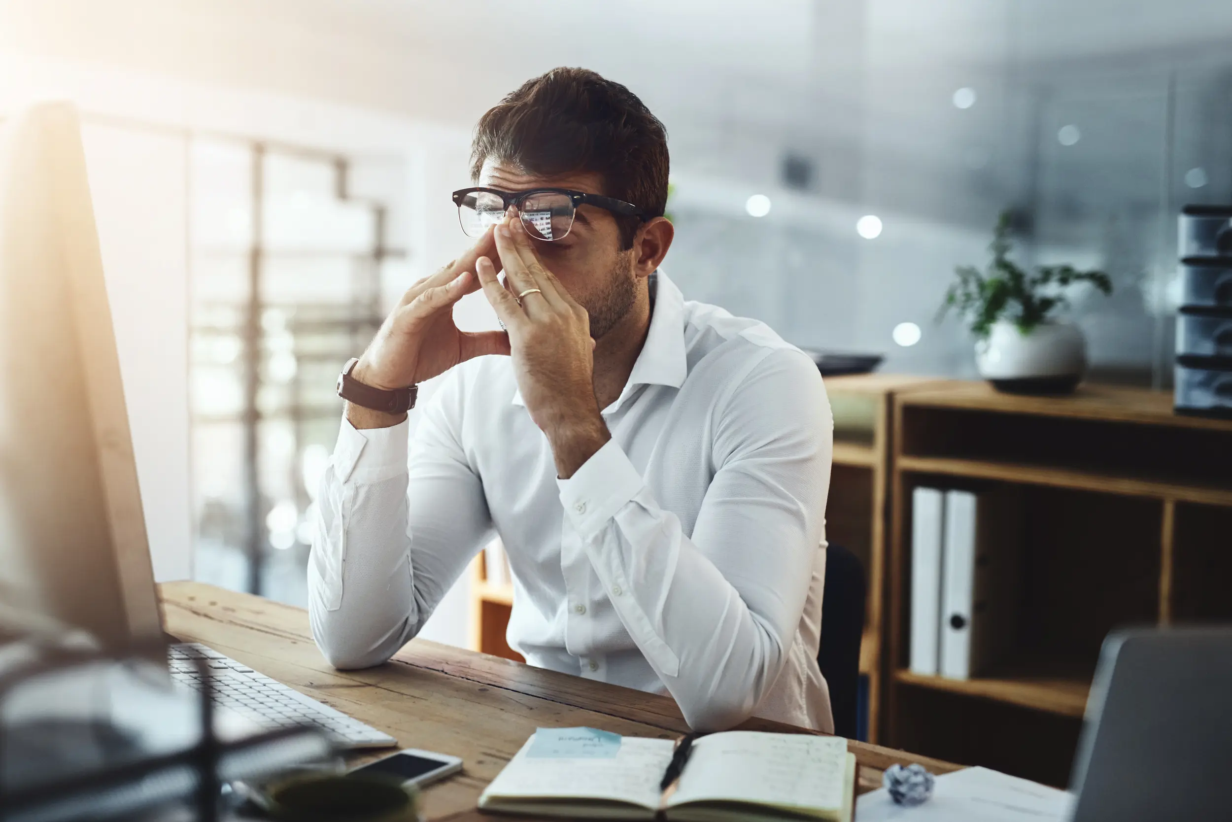 Homem de óculos com expressão cansada e estressada em escritório, representando as profissões que causam infelicidade no trabalho.