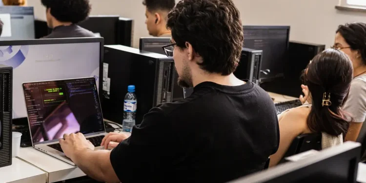 Jovens estudantes em laboratório de informática com notebooks e computadores realizando atividades práticas de cibersegurança no programa Hackers do Bem, iniciativa gratuita da Rede Nacional de Ensino e Pesquisa com mais de 25 mil vagas abertas.