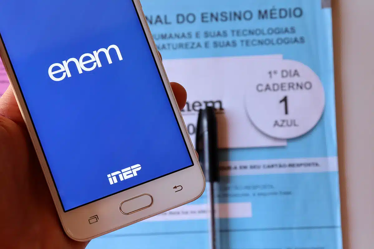 Mão segurando smartphone com o aplicativo do Enem aberto na tela, ao lado do caderno de questões oficial do Enem e uma caneta, representando a preparação digital para a prova de Língua Portuguesa do Enem 2026.