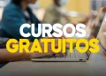 Mãos digitando em um notebook em ambiente de estudo com outras pessoas ao fundo desfocadas e a inscrição "Cursos Gratuitos" em letras brancas e amarelas em destaque, representando o acesso à plataforma QualificaPro lançada pelo governo federal para capacitar trabalhadores