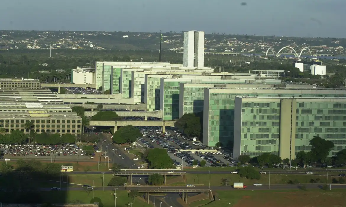 Vista aérea da Esplanada dos Ministérios em Brasília, mostrando os blocos ministeriais alinhados, gramados verdes, estacionamentos e o Congresso Nacional ao fundo sob luz solar.