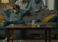 Em um ambiente doméstico com luz suave à noite, um adolescente do sexo masculino, com uma blusa listrada de azul e branco, está sentado em um sofá cinza com as mãos na cabeça e expressão de angústia. Ao fundo, seus pais idosos, um homem e uma mulher de cabelos brancos, o observam com preocupação e tentativa de diálogo, mas ele parece isolado em seu próprio mundo. Na mesa de centro, há objetos como um controle remoto e livros, simbolizando a desconexão familiar diante da tecnologia.
